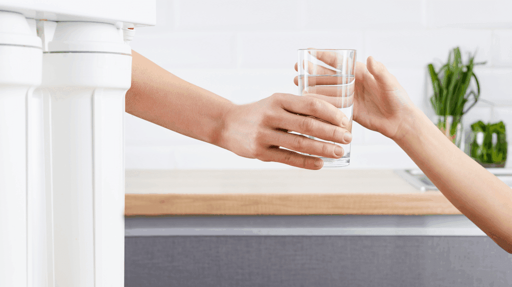AWA Purification Technologies water filtration systems South Texas - A person hands a clear glass of water to another in a modern residential kitchen, featuring a white countertop and green plants, highlighting the benefits of commercial water purification systems.