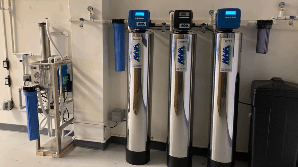 AWA Purification Technologies water filtration systems South Texas - Three large metallic water purification systems with digital monitors stand beside a smaller commercial filtration unit mounted on the wall in a clean, white room with visible pipes and a black container.